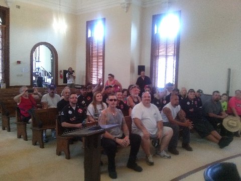 A group of people, including several men wearing black shirts with KKK insignia, are seated in wooden pews inside a large room with arched windows, possibly a courtroom.