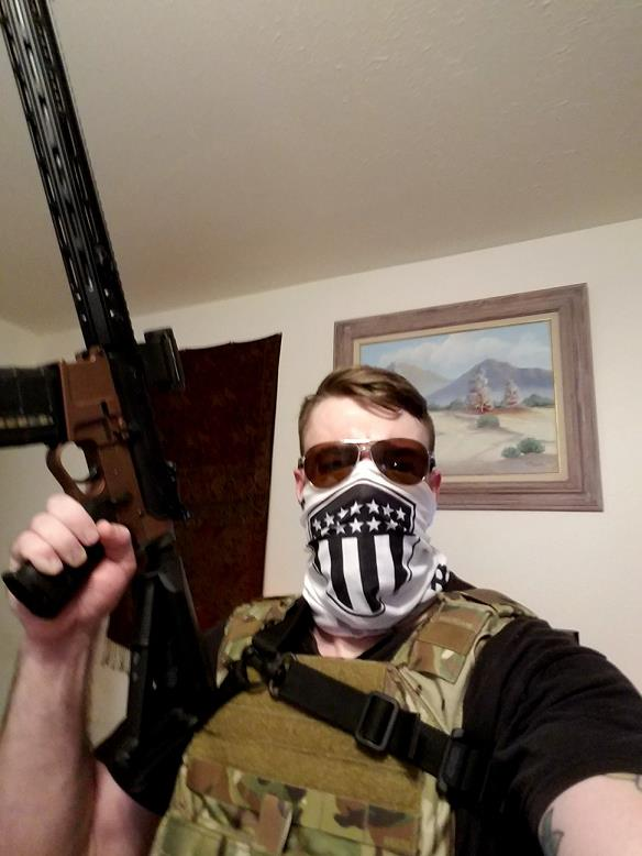 A man wearing a tactical vest, sunglasses, and a face mask with a shield emblem, holding a rifle.