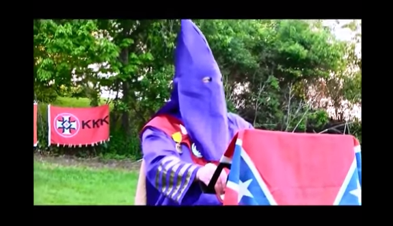 A person in a purple Ku Klux Klan robe and hood stands behind a podium draped with a Confederate flag, with a KKK flag in the background.