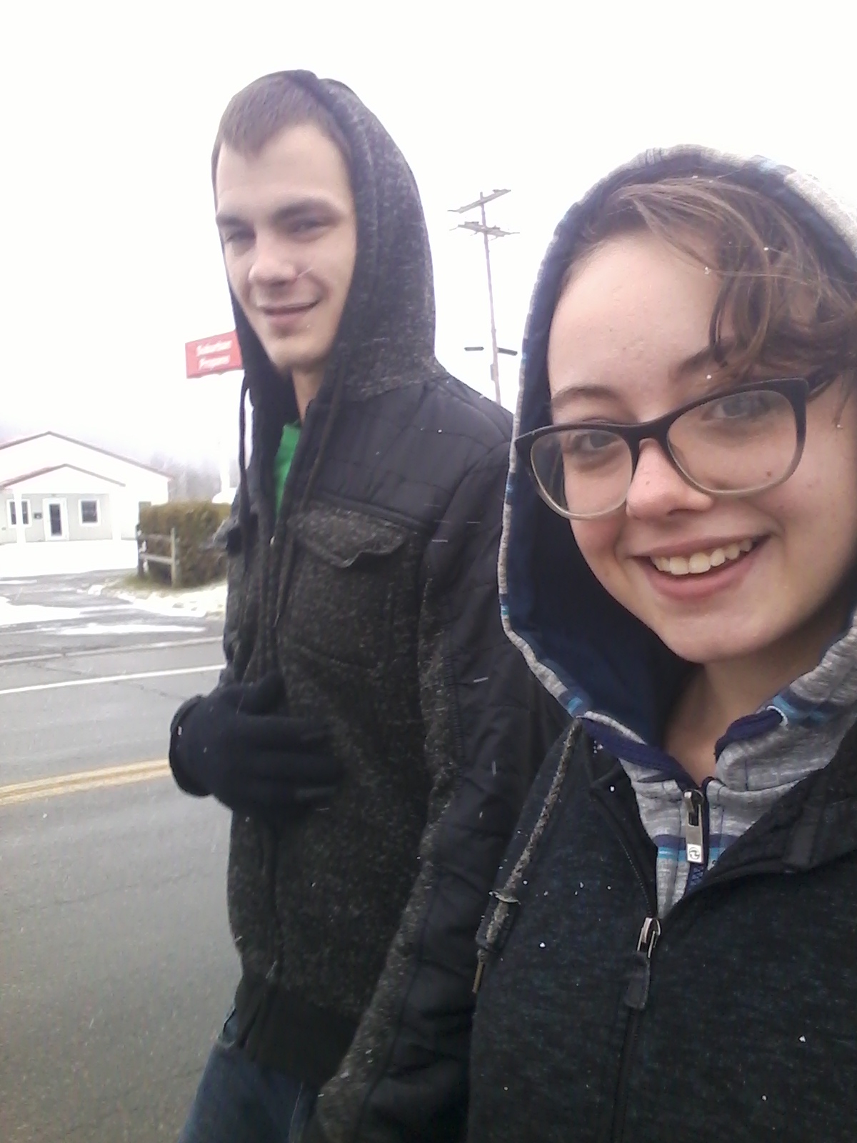 A selfie of a young man and a young woman wearing hooded jackets, walking along a road in a snowy or overcast environment.