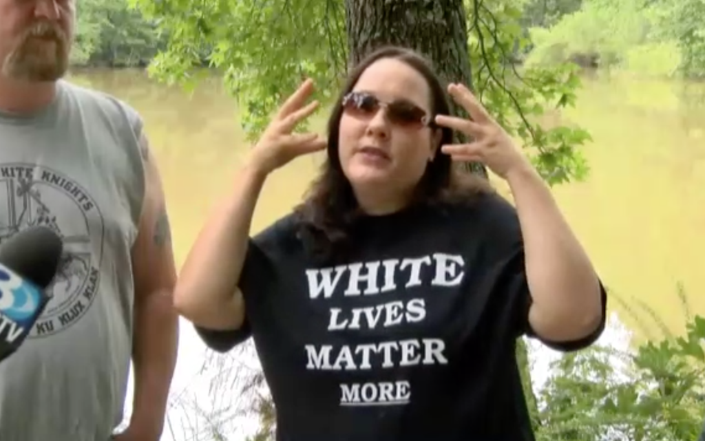 A woman wearing a black t-shirt that reads 'WHITE LIVES MATTER MORE' is being interviewed outdoors near a river. To her left, a man with a goatee and a grey t-shirt featuring a 'Loyal White Knights Ku Klux Klan' logo is partially visible.