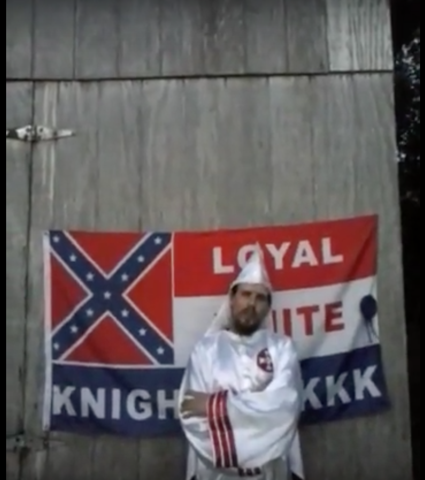 A man wearing white Ku Klux Klan robes and a pointed hood stands with arms crossed in front of a 'Loyal White Knights KKK' flag pinned to a wooden wall.