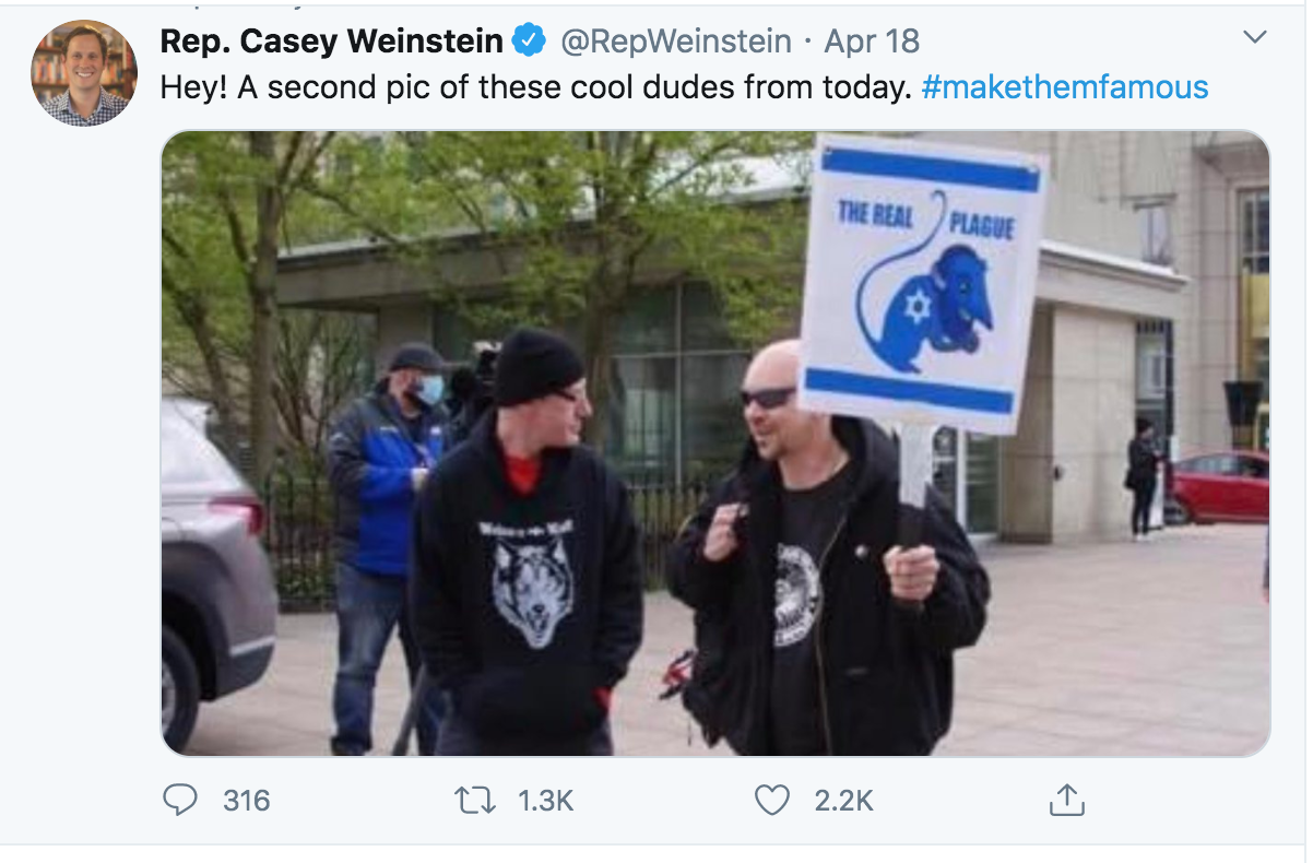 A screenshot of a tweet from Rep. Casey Weinstein showing two men at a protest. The man on the right is bald, wearing sunglasses and a black jacket, holding a sign with an antisemitic caricature and the text 'THE REAL PLAGUE'. The man on the left wears a black beanie and a hoodie with a wolf logo.