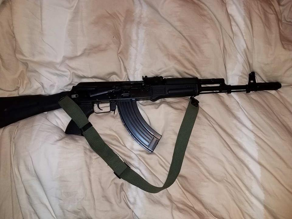 A black AK-style rifle with a green sling resting on a white bedsheet.