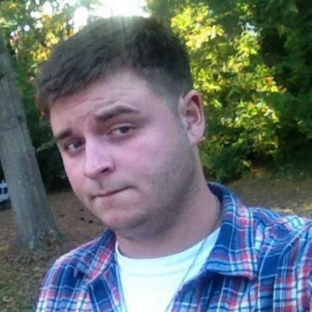 A close-up photograph of a young Caucasian man with short dark hair, wearing a blue and red plaid shirt over a white t-shirt, standing outdoors with trees in the background.