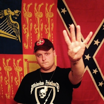 A young Caucasian man wearing a black Phinehas Priesthood t-shirt and a baseball cap with a KKK insignia, holding up four fingers. He stands in front of a Confederate battle flag and a red and yellow heraldic flag.
