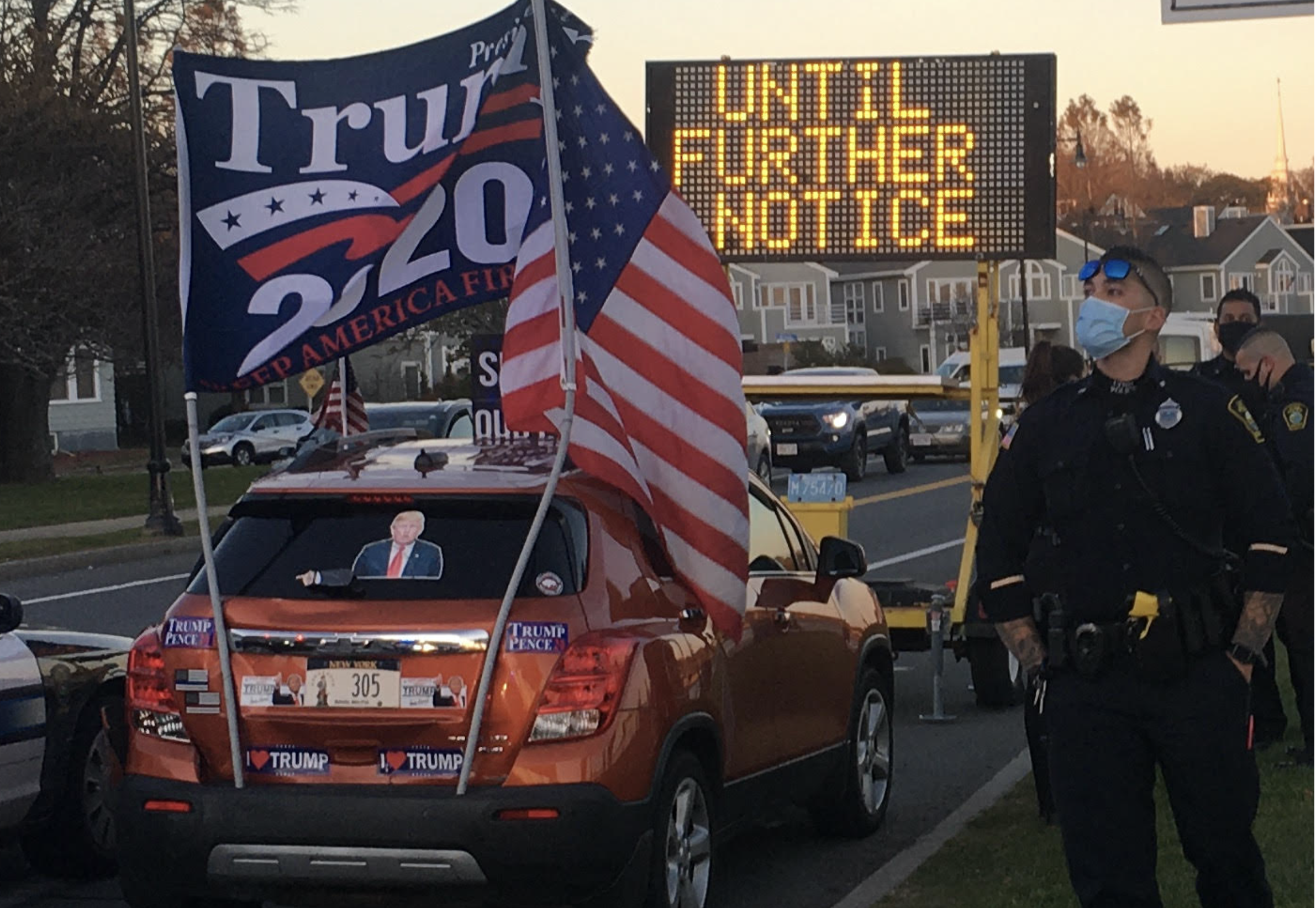 An orange SUV decorated with Trump 2020 flags and stickers is parked on a street next to a digital road sign that reads 'UNTIL FURTHER NOTICE'. A police officer wearing a face mask stands in the foreground.