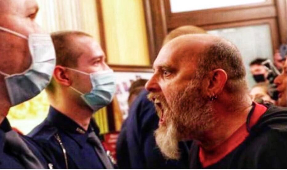 A photograph showing a man with a balding head and a thick, full beard shouting aggressively at police officers who are wearing surgical face masks.