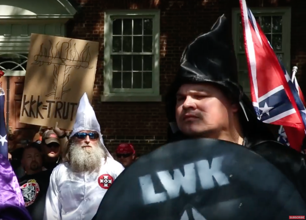 A photograph of a KKK rally featuring individuals in traditional white and black robes and hoods. One person holds a shield with 'LWK' (Loyal White Knights) written on it, while another holds a sign that reads 'KKK=TRUTH'. A Confederate flag is visible in the background.