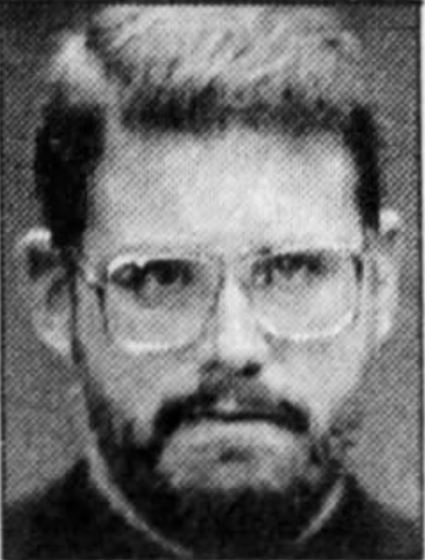 A grainy black and white headshot of a man with a full head of hair, a beard, and large glasses.
