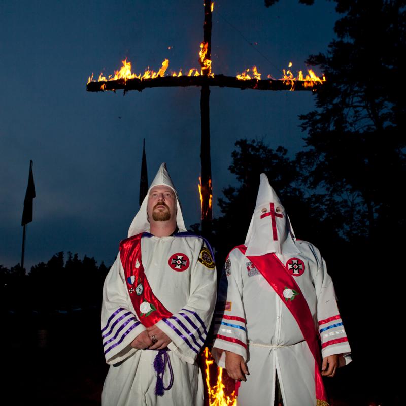 Two individuals in white Ku Klux Klan robes and red sashes stand in front of a large burning cross at night. One man has his hood pulled back, revealing his face and a goatee.