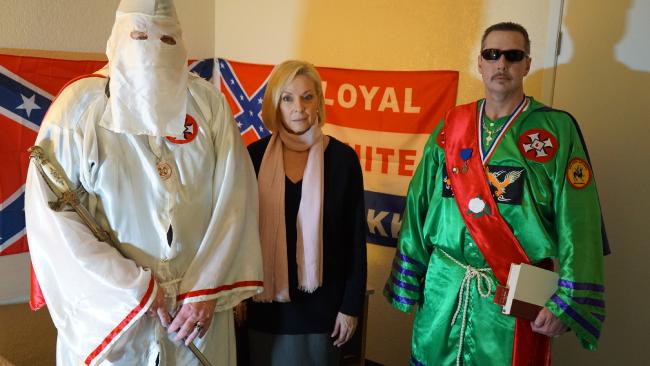 Three individuals standing in front of Confederate and Loyal White Knights flags. One person is in a white KKK robe and hood holding a sword, a woman stands in the center, and a man in a green robe with a red sash and sunglasses stands on the right.