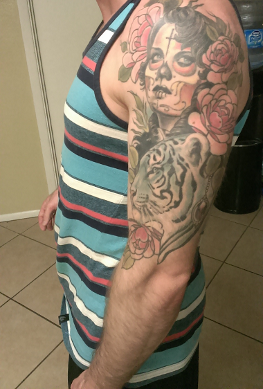 A side-view photograph of a man's upper arm and torso. He is wearing a blue, white, and red striped tank top. His arm features a large, colorful tattoo depicting a woman with sugar skull makeup, roses, and a tiger's head.