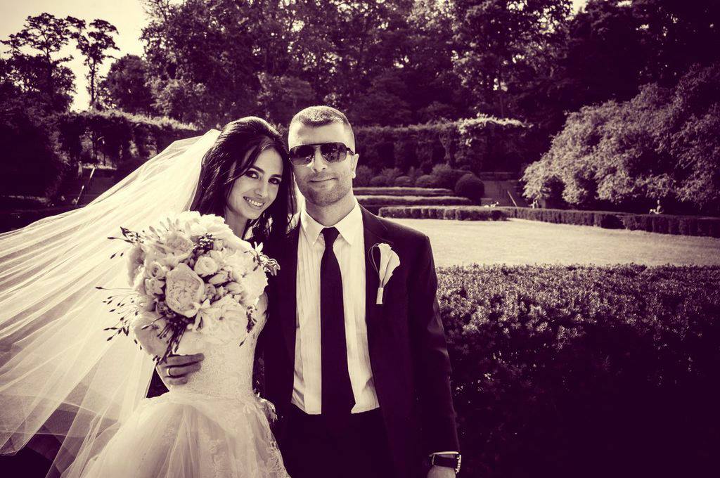A sepia-toned photograph of a bride and groom posing in a formal garden setting. The bride wears a white gown and veil, while the groom wears a dark suit, tie, and sunglasses.
