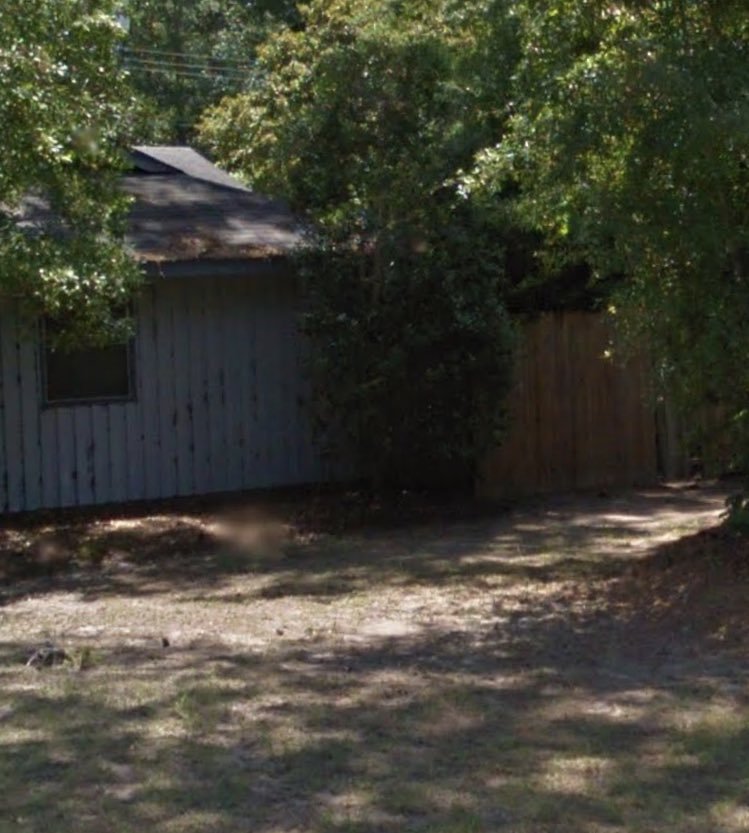 A photograph of a weathered grey shed or small building partially obscured by trees and a wooden fence.