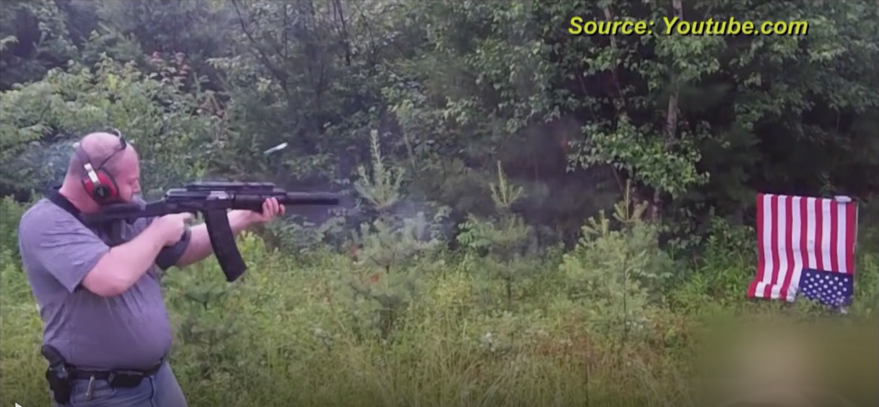 A screenshot of a heavy-set bald Caucasian man in a grey t-shirt firing a rifle in a wooded area. He is wearing red ear protection and has a handgun holstered. An upside-down American flag is in the background.