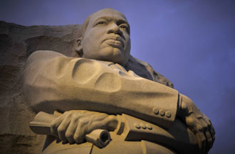 A low-angle photograph of the Martin Luther King Jr. Memorial sculpture in Washington, D.C., showing the figure carved from light-colored stone against a dark blue sky.