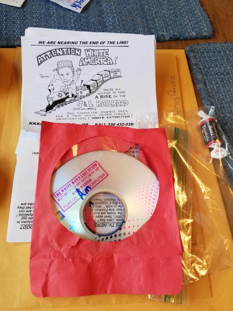 A photograph of KKK propaganda materials including a flyer with a racist cartoon titled 'ATTENTION WHITE AMERICA!', a CD titled 'The White Man's Last Stand' in a red sleeve, and a Tootsie Roll candy.