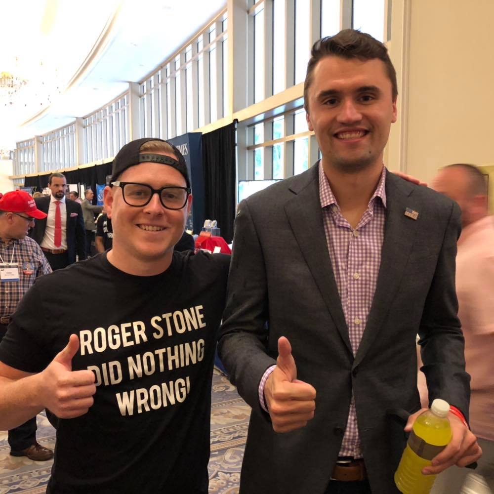 A photograph of two men posing at a political event. The man on the left wears a black t-shirt with the text 'ROGER STONE DID NOTHING WRONG!', a backwards black cap, and glasses. The man on the right, Charlie Kirk, wears a grey suit jacket and a checkered shirt. Both are giving a thumbs-up gesture.