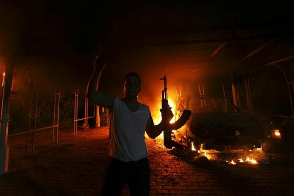 A man in a white tank top holds a rifle aloft in front of a burning car at night during a riot or attack.