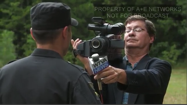 A screenshot from an A+E Networks production showing a man with brown hair and glasses holding a professional video camera and a WTVA News microphone while filming a person in a black uniform.