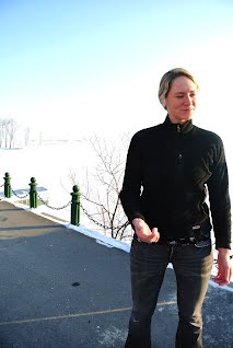 A photograph of a young woman with blonde hair wearing a black zip-up jacket and jeans, standing outdoors in a snowy landscape next to a chain-link fence.