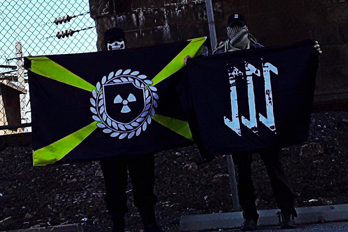 Two masked individuals standing outdoors holding two flags. The flag on the left features a yellow X on a black background with a central emblem containing a radiation symbol inside a shield surrounded by a wreath. The flag on the right is black with three white stylized arrows pointing downwards.