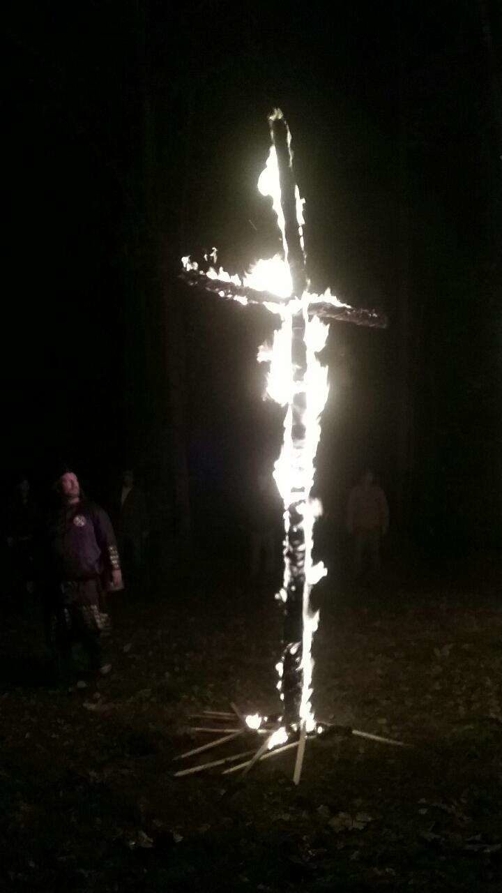 A large wooden cross burning at night in a wooded area, with several indistinct figures standing in the background.