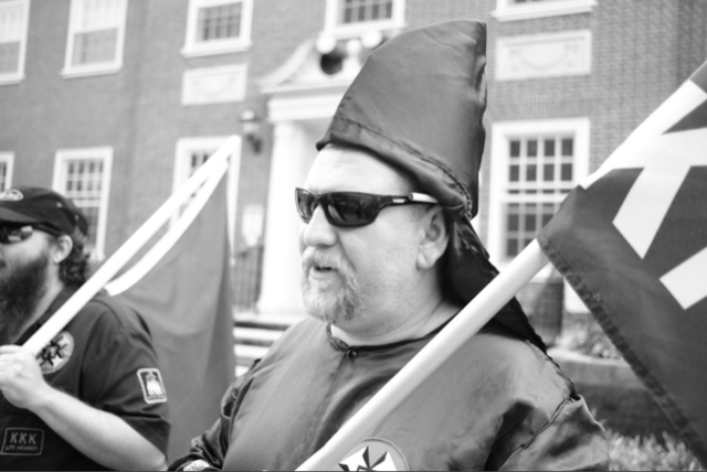 A black and white photograph of a man in KKK regalia, including a pointed hood and robe, wearing sunglasses and sporting a goatee. He is holding a flagpole. In the background, another man with a full beard and a baseball cap is partially visible.