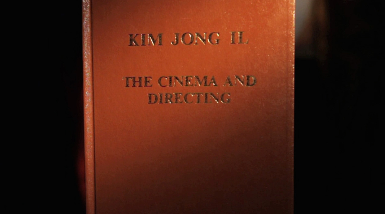 A close-up photograph of a book cover titled 'KIM JONG IL: THE CINEMA AND DIRECTING' in gold lettering on a reddish-brown surface.