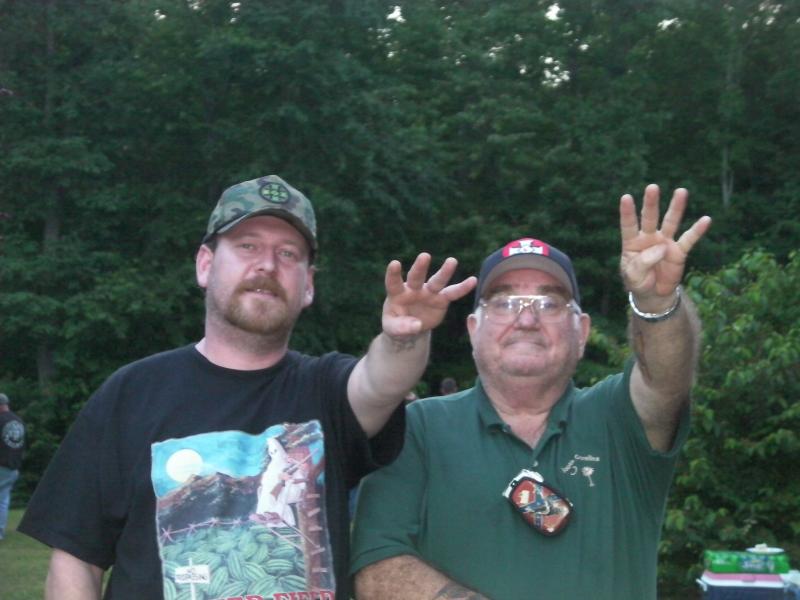 Two Caucasian men standing outdoors in front of a wooded background. The man on the left wears a camouflage hat with a Celtic cross and a black t-shirt featuring KKK imagery. The man on the right wears a green polo shirt and glasses. Both men are holding up four fingers.