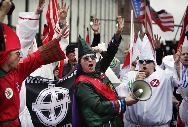 A group of individuals in various colored Ku Klux Klan robes, including red, green, and white, are seen at a rally. A man in a green robe and sunglasses shouts into a megaphone while others raise their hands. A Confederate flag and a banner with a Celtic cross are visible in the background.