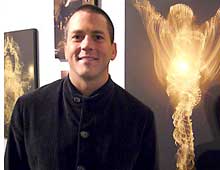 A photograph of a man with short-cropped hair wearing a dark jacket, standing in front of his artwork.