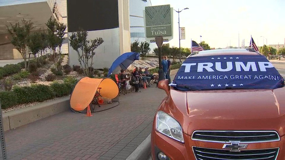A street scene in Downtown Tulsa featuring an orange Chevrolet car with a 'TRUMP MAKE AMERICA GREAT AGAIN!' banner over the windshield. In the background, a small group of people are sitting in chairs under a blue umbrella and an orange pop-up tent on the sidewalk.