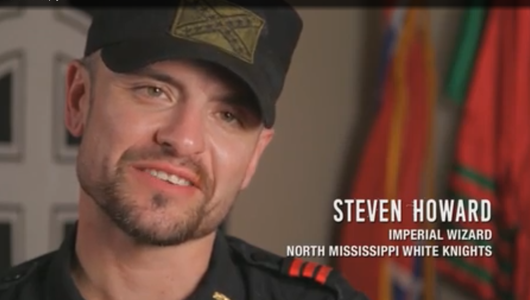 A video still of Steven Howard, identified by on-screen text as the Imperial Wizard of the North Mississippi White Knights, wearing a black cap with a Confederate flag patch and a black uniform shirt.