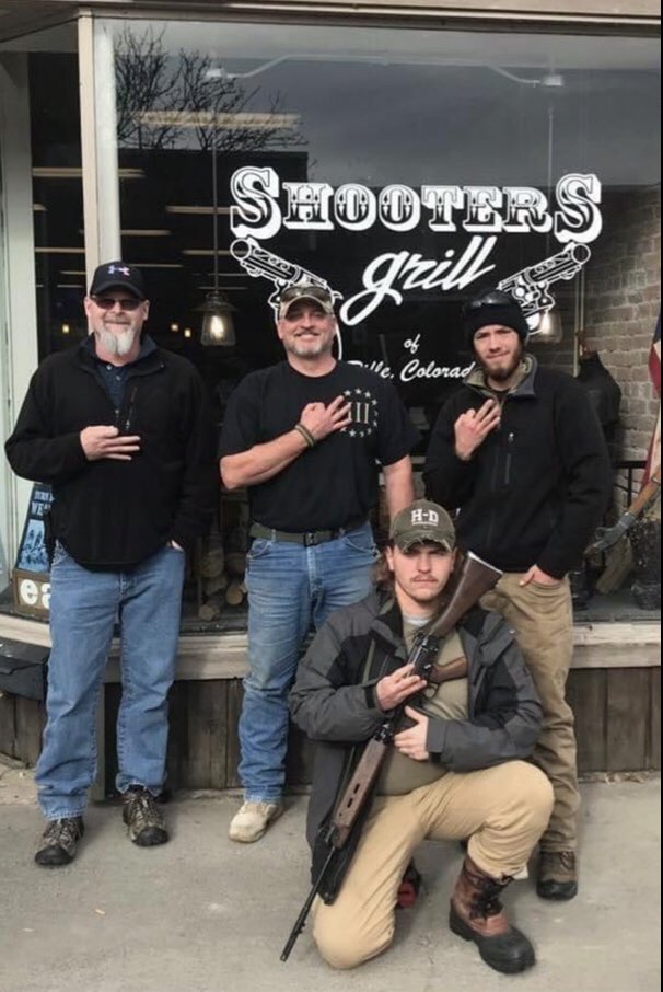 Four Caucasian men posing in front of 'SHOOTERS Grill'. Three men are making hand gestures, and one man is crouching while holding a rifle. The men are wearing casual clothing and hats.