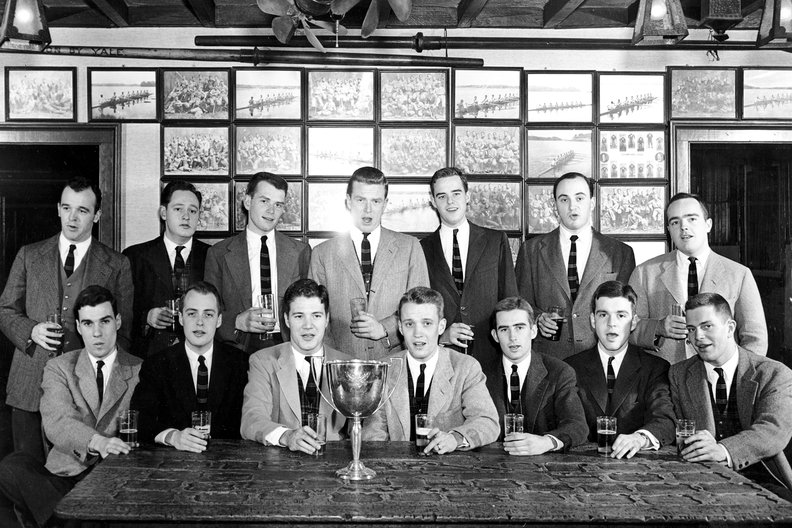 A black and white group photograph of fourteen young men in suits and ties, posed around a table with a trophy and holding glasses. The background features a wall covered in framed photos of rowing teams.
