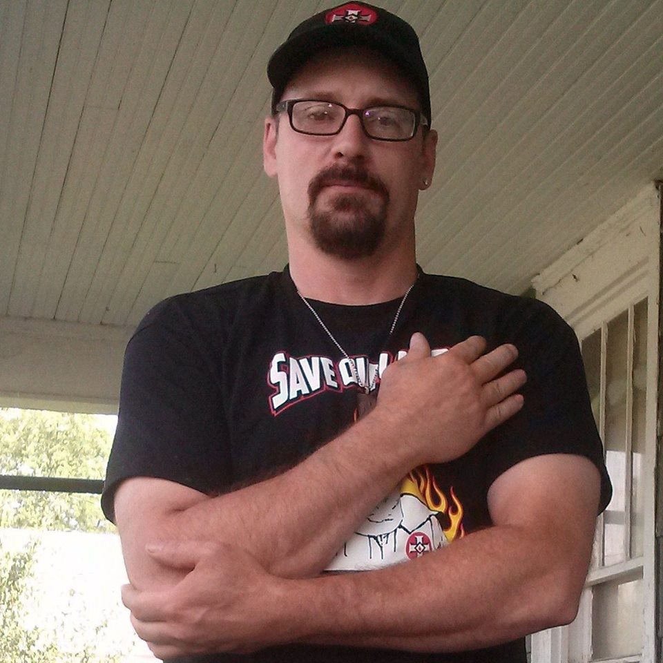 A photograph of a Caucasian man with a goatee and glasses, wearing a black baseball cap with a KKK-style cross symbol and a black t-shirt with white supremacist graphics. He is standing on a porch with his arms crossed.