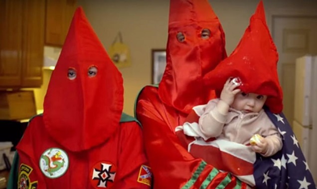 A photograph showing two individuals in red Ku Klux Klan robes and pointed hoods, with one holding a baby also wearing a small red hood and wrapped in an American flag.