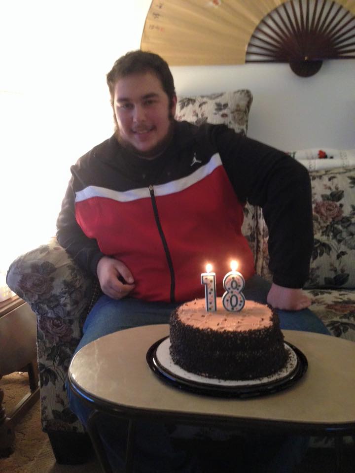A young Caucasian man with dark hair and a beard sits behind a birthday cake with candles showing the number 18.
